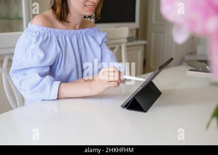 Young brunette woman designer in stylish blouse develops new clothes via tablet on stand at white table near flowers at home close view Stock Photo