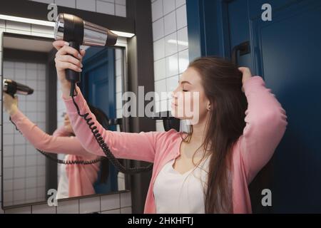 Gorgeous young woman blow drying her hair in hostel bathroom Stock Photo