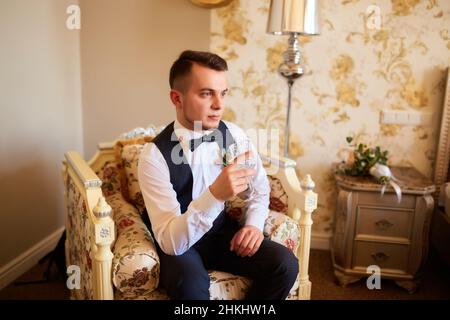 Morning of groom. Beautiful man, groom posing and preparing for wedding. Stylish groom sits on the black chair in studio and holds a glass of whiskey. Stock Photo