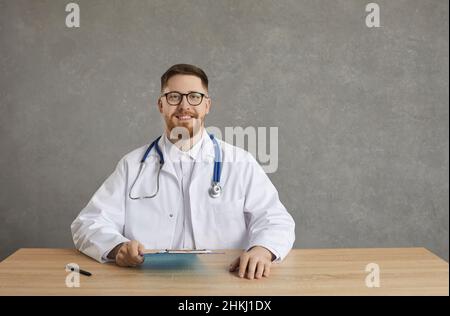 Portrait of confident joyful and smiling male doctor sitting at table on gray concrete background. Stock Photo