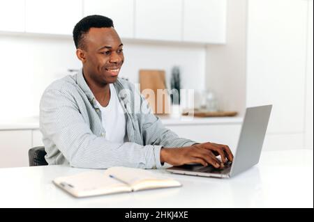 Attractive positive african american guy, freelancer, designer, IT specialist, works on a laptop at home, develops a strategy, analyzes, messaging with clients or colleagues, browsing internet, smiles Stock Photo