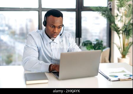 Smart purposeful, focused African American young man in headphones, manager, support worker, online consultant, sitting at a laptop in the office, having an online consultation with clients in a chat Stock Photo