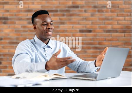 Online business conference. A young positive black guy in a shirt, the company's CEO, manager, broker, trader is having a video call in office with colleagues or partners to discuss the work strategy Stock Photo