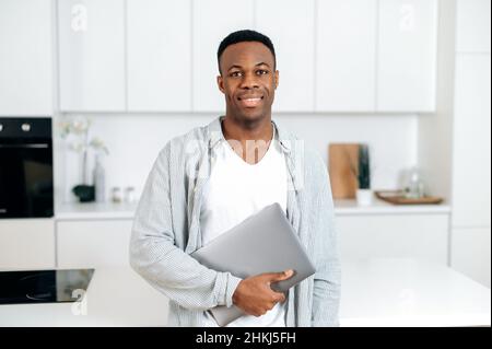 Portrait African American handsome confident guy, freelancer or IT specialist, standing at home in the kitchen, dressed in casual clothes, holding laptop, looking at camera and smiling friendly Stock Photo