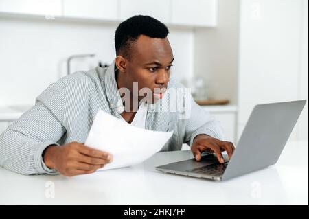 Concentrated confused young african american man, freelancer, stock trader, sitting at the table at home, in casual wear, in confusion looks into a laptop, analyzes online reporting, profit Stock Photo