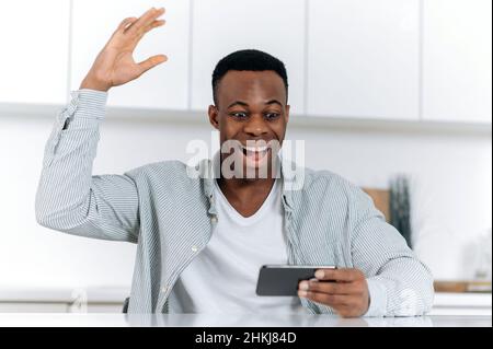 Young adult positive joyful successful african american man sitting at the table with a phone in his hands, rejoicing in victory, news or message, gesturing with hand, looking at the screen, smiling Stock Photo