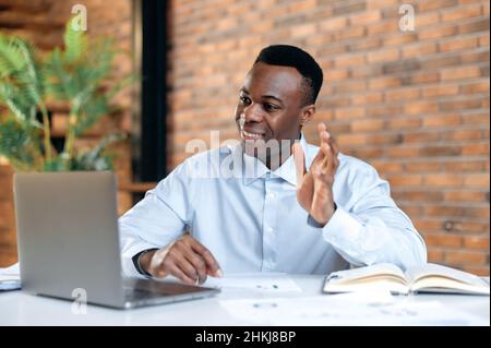 Pleasant friendly black guy, manager or financial consultant, communicates with colleagues on a video call, discusses results and strategy in a modern office, remote negotiations, greeting gesture Stock Photo