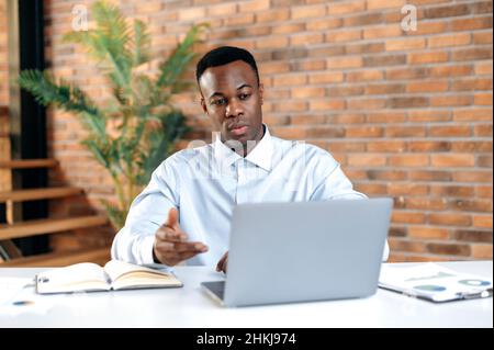 Serious puzzled young adult african american businessman, CEO or top manager sits in modern office, uses a laptop, for an online video call, presentation a business strategy or report, distant talking Stock Photo