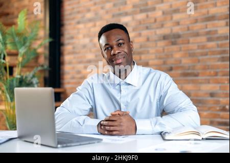 Pleasant young afro american man working on a computer in a modern office. Portrait of a successful businessman, freelancer, manager, wearing a formal shirt, looking at the camera, smiling friendly. Stock Photo