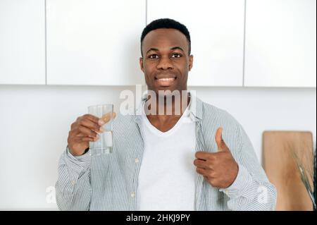 Happy african american young healthy man, standing in the kitchen, in casual clothes, holds a glass of clean water in his hand, looks at the camera, smiles, care about health, shows thumbs up gesture Stock Photo