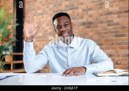 Close-up of webcam headshot, friendly, smart young black male, top manager, financial advisor, sitting at the table in office, talking with colleagues, participating in video call, waving, smiling Stock Photo