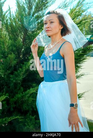 A young beautiful girl in a veil, a white skirt, and blue T-shirt near a juniper bush. The girl is waiting for someone. Stock Photo