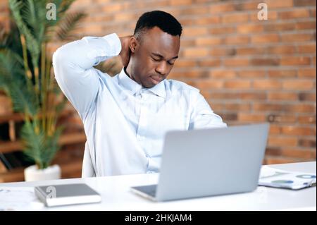 Exhausted by online work african american businessman, sales manager, trader sitting at a table in office, massaging his neck, overworked from negotiations, computer work, in need of rest or break Stock Photo