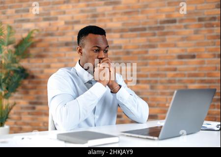 Tense focused young adult african american businessman, CEO or broker, sitting in office at a laptop, works, analyzes strategies, develops a work plan, keeps half a profit and loss of the company Stock Photo