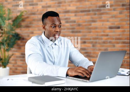 Shocked surprised African American business man, secretary, office worker, sitting at his desk in the office, looking at laptop in amazement, getting unexpected news, winning or making big profit. Stock Photo