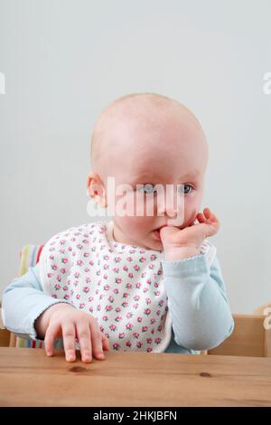 Baby girl sitting at table with a finger in her mouth Stock Photo