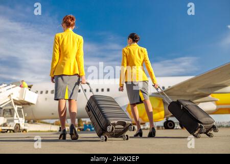 Back view of two women flight attendants carrying trolley luggage bags while walking down airfield Stock Photo