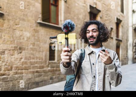 Glad bearded male blogger recording video on cellphone with microphone while standing near shabby stone building in city during trip Stock Photo