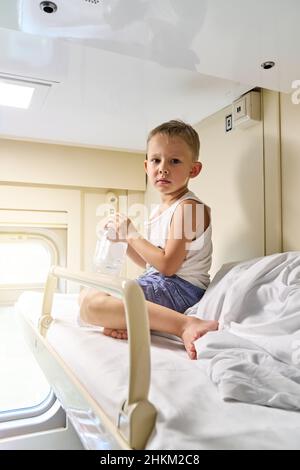 Blond short-haired little boy in t-shirt and shorts sitting on top shelf in train car against bright sunlight closeup Stock Photo