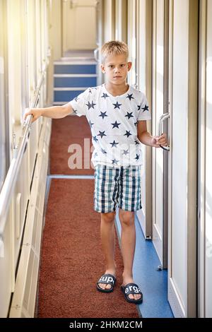 Boy with short blond hair wearing casual clothes opens door into train compartment entering coupe from corridor with bright sunlight Stock Photo