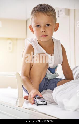 Blond short-haired little boy in t-shirt and shorts plays with car toy sitting on top shelf in train car against bright sunlight closeup Stock Photo