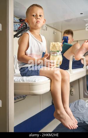 Brothers younger and elder sit on top shelf of train car playing games using smartphone and eating snacks from package closeup Stock Photo