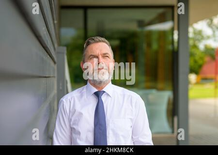 Senior businessman walking through town deep in thought looking up with a contemplative expression as he approaches the camera along a covered walkway Stock Photo