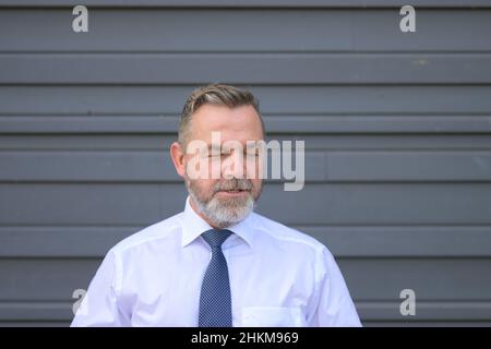Senior bearded businessman speaking to the camera with open mouth and closed eyes over a grey wall background Stock Photo