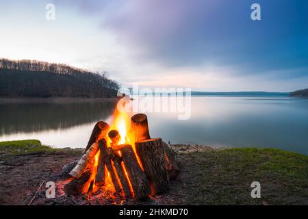 Big campfire with flames beside a lake during a winter sunset. Stock Photo