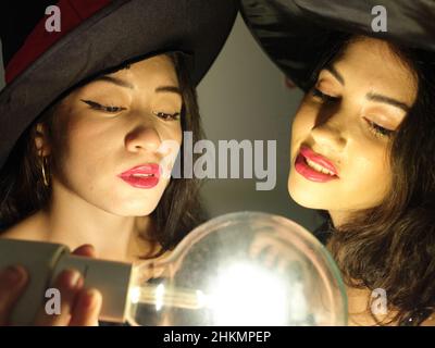 Two latina friends dressed as witches on halloween with a light bulb near them and looking at it. Stock Photo