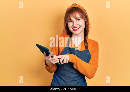 Redhead young woman wearing waitress apron holding touchpad device taking order looking positive and happy standing and smiling with a confident smile Stock Photo