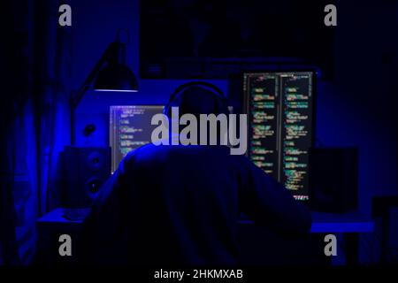 A man in hoodie sits at a computer in a room at a table at night with blue lighting and programs Stock Photo