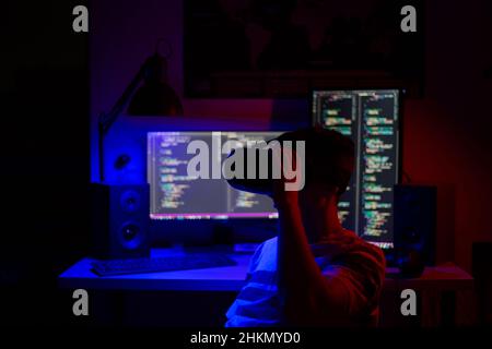A man in VR glasses sits at a computer in a room at a table at night with blue lighting and programs Stock Photo
