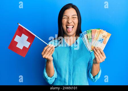 Young latin woman holding switzerland flag and franc banknotes sticking tongue out happy with funny expression. Stock Photo