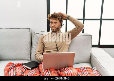 Young handsome man with beard wearing operator headset working from home confuse and wondering about question. uncertain with doubt, thinking with han Stock Photo
