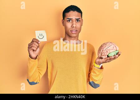 Young african american guy holding smile reminder and brain in shock face, looking skeptical and sarcastic, surprised with open mouth Stock Photo