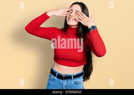 Young brunette teenager wearing red turtleneck sweater covering eyes with hands smiling cheerful and funny. blind concept. Stock Photo