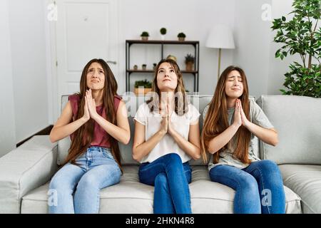Group of three hispanic girls sitting on the sofa at home begging and praying with hands together with hope expression on face very emotional and worr Stock Photo
