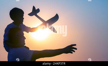 Silhouette of a boy, sitting on his father's shoulder, launches a model airplane into the evening sky against the backdrop of the setting sun. Childre Stock Photo