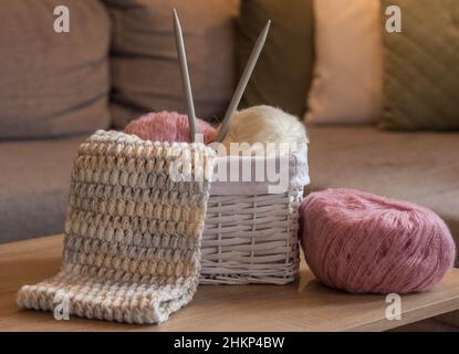 pink and white woolen balls in a white basket with knitting needles and a knitted scarf handmade Stock Photo