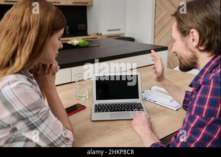 Soft focus on the blank black screen monitor of a laptop with copy space and a happy young couple, man and woman talking by video call sitting at the Stock Photo
