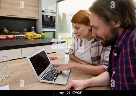 Happy loving Caucasian couple sitting at the kitchen table in front of a laptop and surfing websites, making online purchases, booking, ticketing, enj Stock Photo