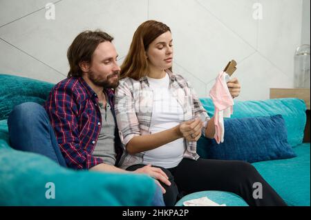 Caucasian loving couple, in anticipation of a baby, rests together sitting on the sofa in an embrace and rejoices, admiring the clothes for the newbor Stock Photo