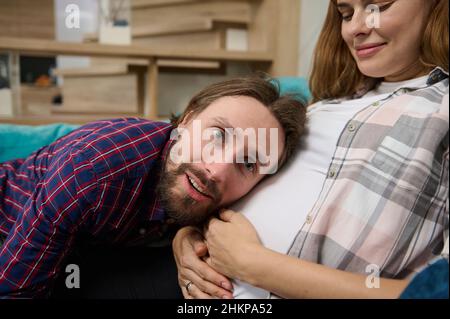 Lovely cute couple, young dad-to-be leaning gently on the belly of his cute pregnant wife, enjoying the expectation of the baby together. Happy and ca Stock Photo