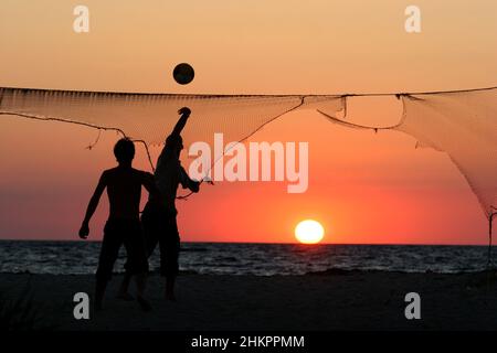 Dark unrecognisable silhouettes of two men playing volleyball over torn net on a beach facing beautiful sunset. Sports activity on a beach Stock Photo