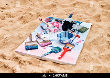 Two painted canvas in blue and pink colors with palettes of paints lying on sandy beach, artist's palette at outdoor art festival. White drawing canva Stock Photo