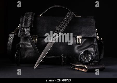 Large folding knife and black leather bag. Knife and tactical watch. Black objects on a black background. Front view. Stock Photo