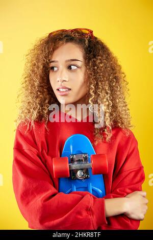 Delighted African American young female hipster in hoodie and with penny board against yellow background Stock Photo