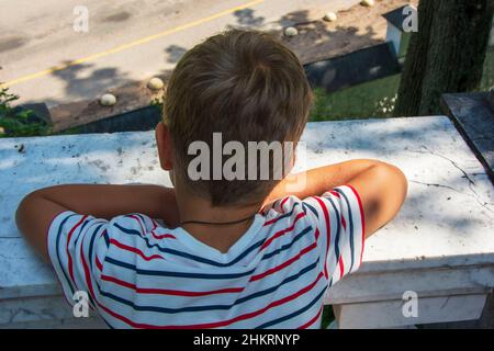 A preschooler looks down from the balcony, rear view. The concept of isolation and loneliness of young children. Stock Photo