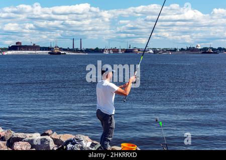 A fisherman with a fishing rod on the shore of the bay against the background of the city, A man holds a fishing rod in his hands. Summer. Stock Photo
